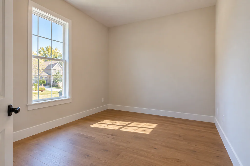 Empty home office with pale walls, wood flooring, and a tall window overlooking the neighborhood.